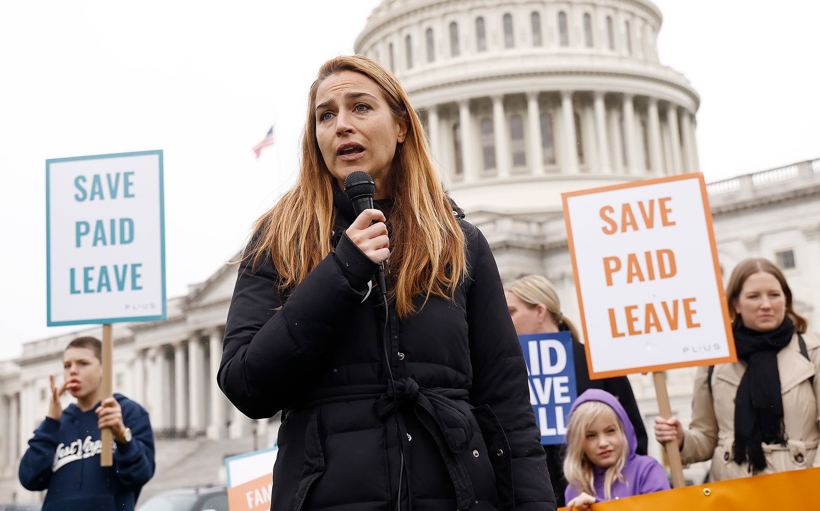 A woman holds a microphone at a rally in front of Congress, surrounded by signs saying 'Save Paid Leave'