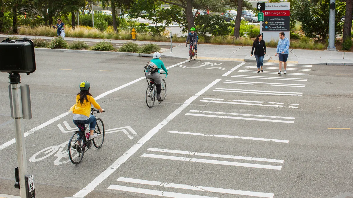 Multiple pedestrians and cyclists cross at an intersection with designated crosswalk lanes. In the background a sign reads, "Seattle Children's Hospital, Emergency, Main Entrance, River Parking.."