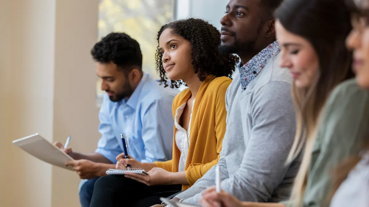 A group of business people sit in a row in a training class. They look at an unseen speaker as they concentrate on his lecture.