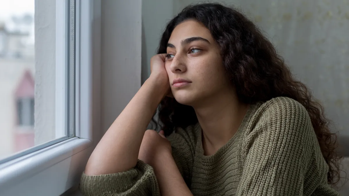 A teenage girl looks sadly out the window with her face on her hand.