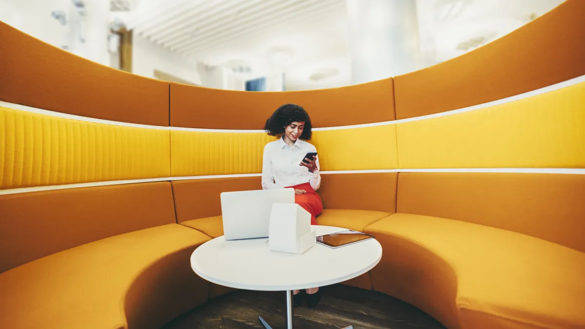 A wide-angle shot of a Black business person using their smartphone while sitting on a round yellow sofa in office coworking area