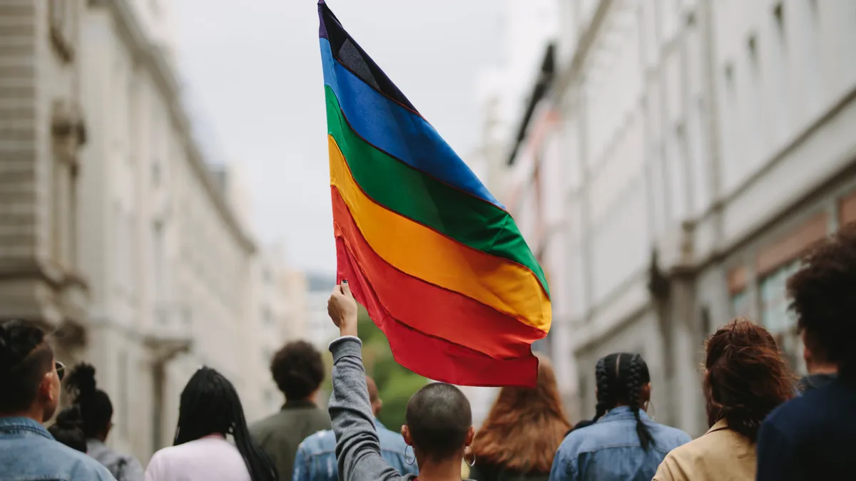 A person waves a large rainbow flag in a small crowd.