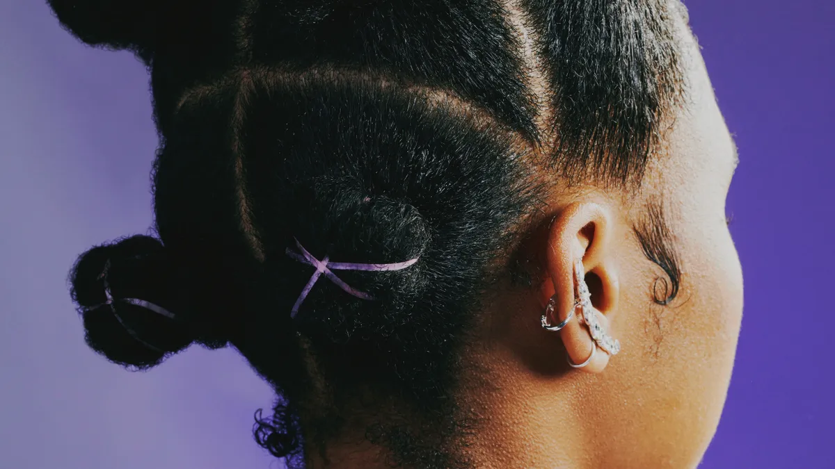 Close up: Bantu knots with purple elastics. A Black woman stands against a purple studio background, wearing a lighter purple windbreak that is zipped up on the back her neck