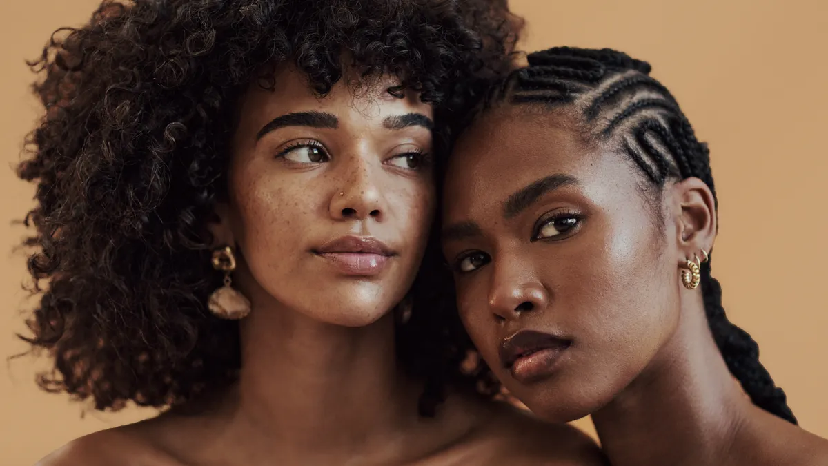 Two Black people — one with an afro, one with braids — pose for a studio portrait with a light brown background
