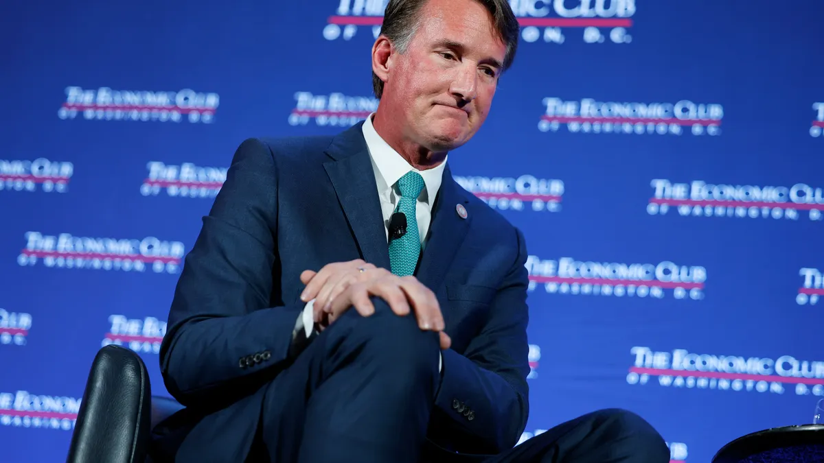 Youngkin sits, mouth slightly downturned with both hands on his right knee in a blue suit and green tie with a microphone. Blue background with The Economic Club of Washington, D.C., in white and red.