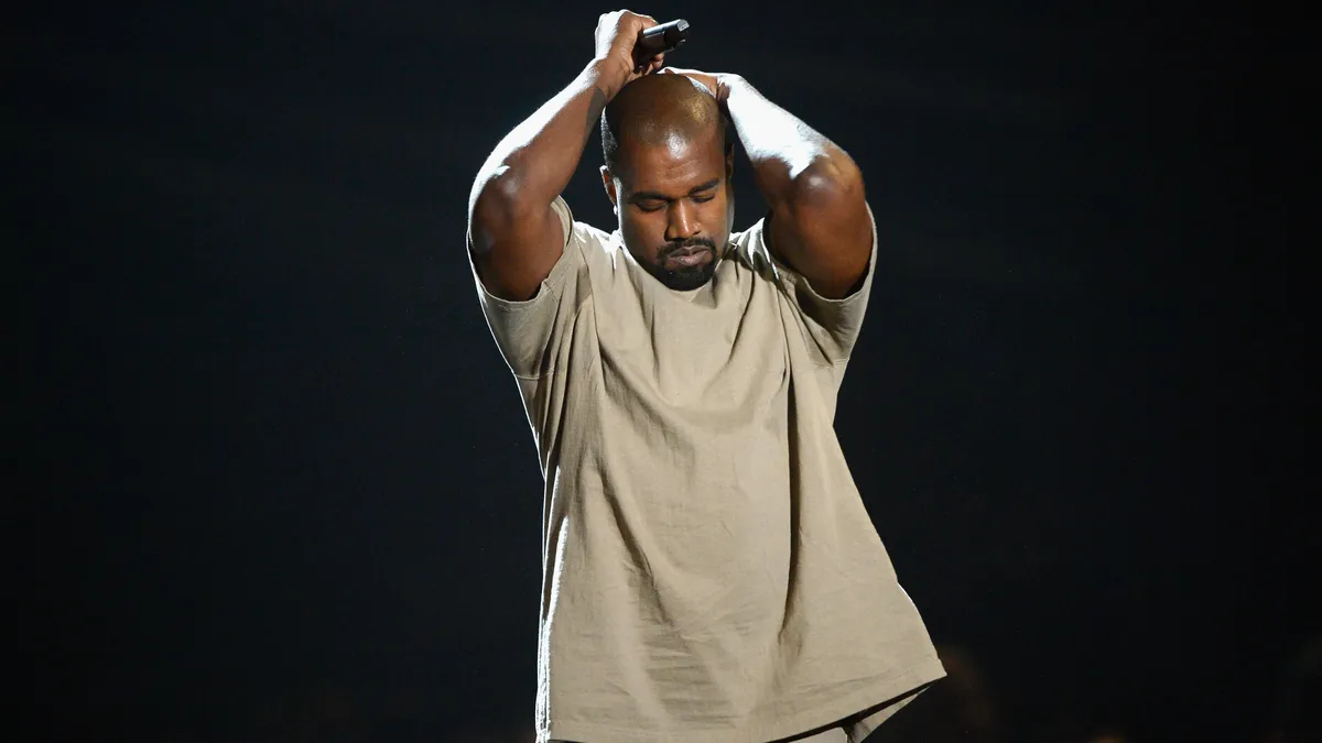 Kanye West onstage during the 2015 MTV Video Music Awards. He is wearing a monochrome ecru outfit and he is clutching his head.