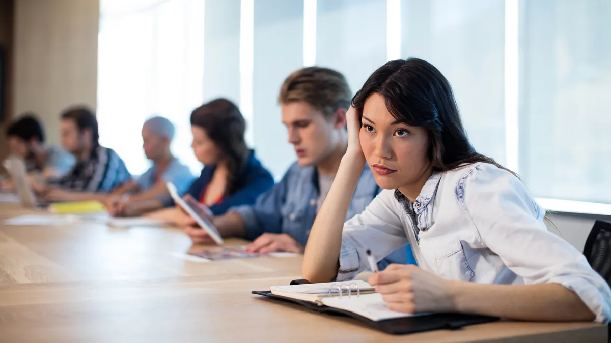 A bored young woman sits in a meeting, with out-of-focus, more engaged workers in the background.