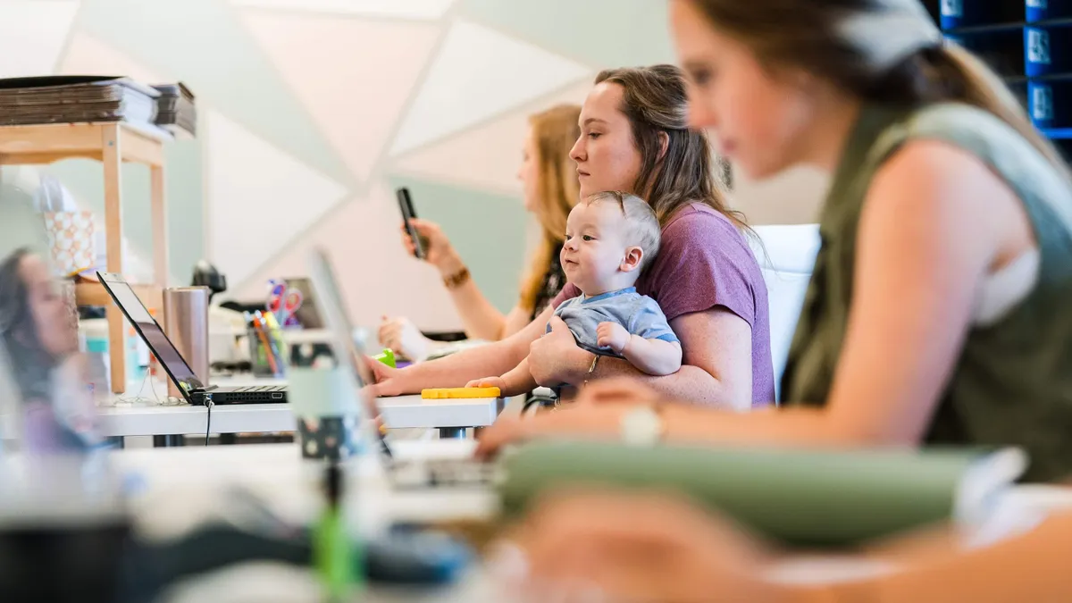 A woman sits with her baby in front of a laptop at a desk.